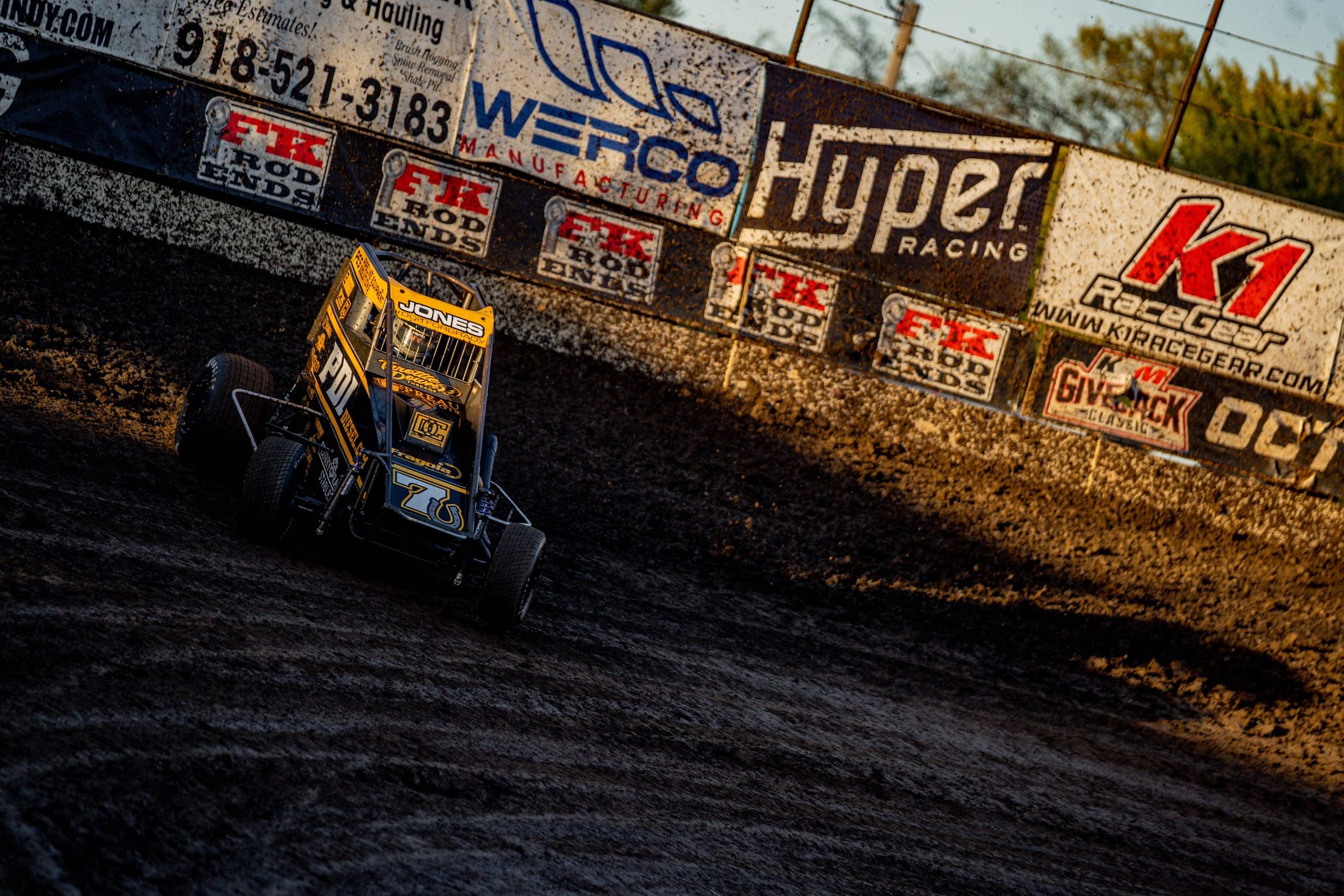 Sprint car sliding past trackside sponsor banners at sunset